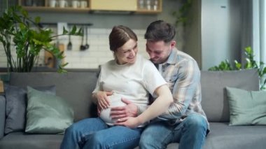 portrait happy family during pregnancy at home. Young happy family Caucasian couple husband and pregnant wife with belly sitting on sofa in living room and looking at camera smiling. Indoor. Happiness