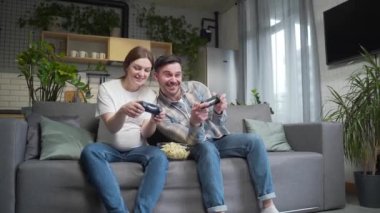 Happy young couple, pregnant wife and husband playing video game with joysticks on console. Cheerful family spending leisure time together during pregnancy sitting together on sofa at home front TV