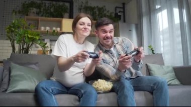 Happy young couple, pregnant wife and husband playing video game with joysticks on console. Cheerful family spending leisure time together during pregnancy sitting together on sofa at home front TV