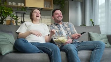 Cheerful family holding remote control laughing watching a hilarious funny TV show. couple husband and pregnant wife at home spending leisure time together on sofa watching comedy video movie enjoy