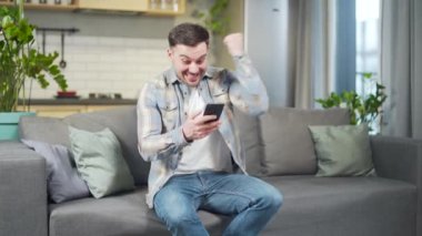 Happy man looking at cellphone rejoices shocked good news, victory in home. Cheerful male look at smartphone mobile phone screen celebrate online winner Surprised Shouts yes sitting on the couch