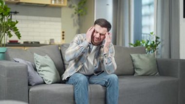 Tired and sick man at home alone, handsome has a headache, sits on the couch, and holds his hands behind his head. portrait of a sick male exhausted massaging the temples. ache, pain, blood pressure