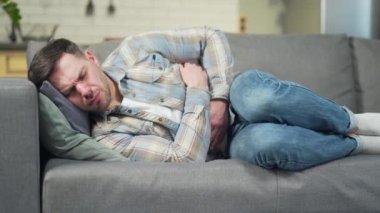 a young handsome man is lying on the sofa at home with severe abdominal pain, stomach upset, diarrhea. Bearded male massaging suffers from gastritis or ulcers. Poisoning, intestinal spasms. indoors