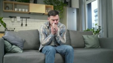 portrait sick handsome bearded man coughing with flu and cold sitting on sofa in living room at home. Caucasian young male having fever runny nose ill suffering disease Feeling unwell