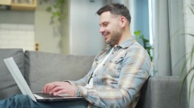 Handsome bearded young man works using a laptop computer. Caucasian male browsing, searching, chatting text message, correspondence sitting on comfortable sofa resting relaxing at home in living room