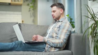 Handsome bearded young man remote meeting via webcam works using laptop computer. Caucasian male talking online via video call sitting on a comfortable sofa chatting resting relaxing at home in room