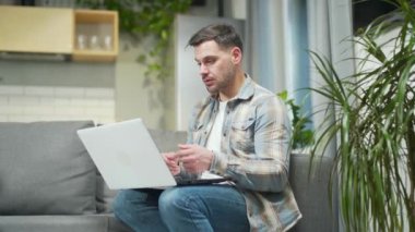 Handsome bearded young man remote meeting via webcam works using laptop computer. Caucasian male talking online via video call sitting on a comfortable sofa chatting resting relaxing at home in room