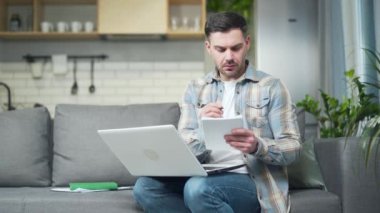 young handsome bearded Caucasian man studying working remotely at home using a laptop computer taking notes in a notebook sitting on the sofa. Online training course or lessons