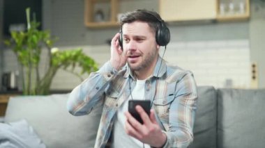 happy young bearded cheerful handsome caucasian man enjoying listening to music sitting at home on sofa in room with headphones using smartphone. The male dances and sings Cheerful rhythmic joyful guy