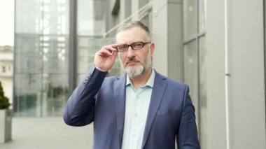 Portrait serious confident business mature man in formal suit looking at camera with crossed arms. Bearded gray-haired Businessman near office building outside stands on the urban street of the city