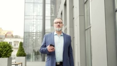 Confident business mature experienced gray-haired man in a formal suit walking down the city street with a smartphone phone in his hands against an urban background of an office building. outside