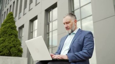 Mature gray-haired business man in formal suit working on laptop online remotely. Sitting on a bench near office building outside. senior employee uses a computer outdoors urban background city street