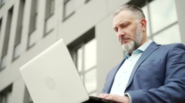 Mature gray-haired business man in formal suit working on laptop online remotely. Sitting on a bench near office building outside. senior employee uses a computer outdoors urban background city street