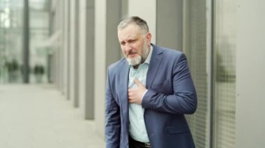 mature business man with a sudden heartache is standing on a city street near office building. Senior businessman, employee holding chest outside outdoor feeling sharp chest pain, symptom heart attack