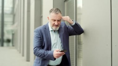 Mature business man with dizziness, vertigo a severe headache standing on a city street leaning against a wall. outside. Senior male with high blood pressure or stroke holding head massaging. Outdoor