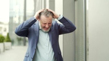 Mature business man with dizziness, vertigo a severe headache standing on a city street leaning against a wall. outside. Senior male with high blood pressure or stroke holding head massaging. Outdoor