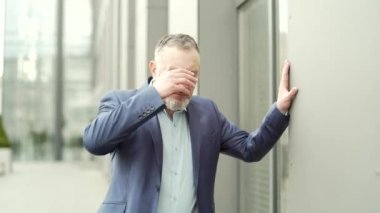 Mature business man with dizziness, vertigo a severe headache standing on a city street leaning against a wall. outside. Senior male with high blood pressure or stroke holding head massaging. Outdoor