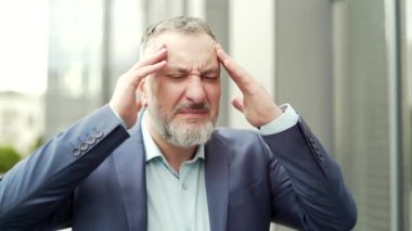 Mature business man with dizziness, vertigo a severe headache standing on a city street leaning against a wall. outside. Senior male with high blood pressure or stroke holding head massaging. Outdoor