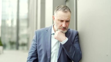 Sad confused mature businessman standing near office building outside. Business man in a suit with depression. Sorrowful male has financial problems. Dismal bored employee entrepreneur on the street