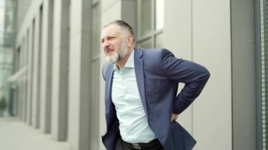 Mature business man holding his back feels back pain at work outside Spine Apin. suffering from backache spinal injury and muscle issue problem outdoor standing on a city street near office building
