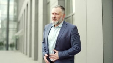 Mature man abdominal pain standing on city street near office building Senior gray-haired businessman outside, male in business suit sick has stomachache. stomach ache nausea, painful disease outdoors