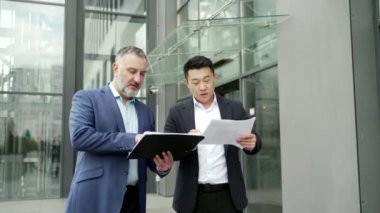Business partners colleagues at work discussing a project while standing near office building. A mature man and an Asian employee in suits talking communicating outside with documents in hands