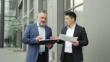 Business partners colleagues at work discussing a project while standing near office building. A mature man and an Asian employee in suits talking communicating outside with documents in hands