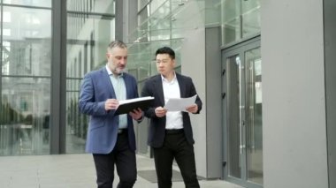 Business partners colleagues at work discussing a project while walking near modern office building. A mature man and an Asian employee in suits talking communicating outside with documents in hands