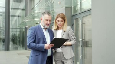 two business partners colleagues at work discussing a project while standing near office building. A boss mature man and woman employee in suits talking communicating outside with documents in hands
