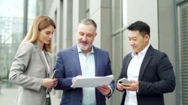 Happy Team Business partners colleagues at work discussing a project stand near modern office building. Mature man, woman, Asian employee in suits talking communicating outside with documents in hands