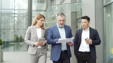 Team Business partners colleagues at work discussing a project walking near modern office building. Mature man, woman and Asian employee in suits talking communicating outside with documents in hands