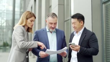Happy Team Business partners colleagues at work discussing a project stand near modern office building. Mature man, woman, Asian employee in suits talking communicating outside with documents in hands