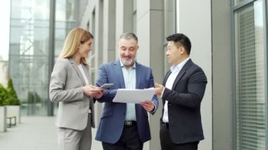 Happy Team Business partners colleagues at work discussing a project stand near modern office building. Mature man, woman, Asian employee in suits talking communicating outside with documents in hands