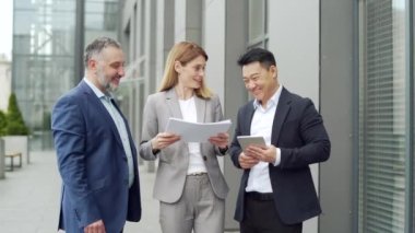 Happy Team Business partners colleagues at work discussing a project stand near modern office building. Mature man, woman, Asian employee in suits talking communicating outside with documents in hands