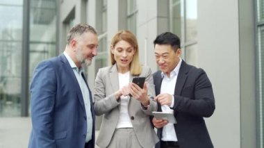group of colleagues fellow employees read good news looking at smartphone. Happy team rejoices about successful investment while using, browsing mobile phone. Standing outside near an office building