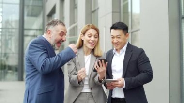 group of colleagues fellow employees read good news looking at smartphone. Happy team rejoices about successful investment while using, browsing mobile phone. Standing outside near an office building