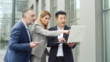 Team of business people employees in formal suits discussing a project together using a laptop and tablet standing near office building. Group partners mature person looking at computer screen outside