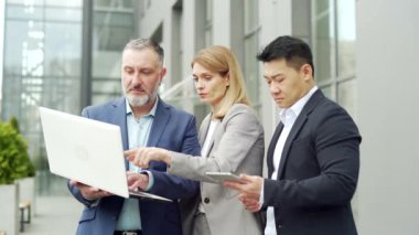 Team of business people employees in formal suits discussing a project together using a laptop and tablet standing near office building. Group partners mature person looking at computer screen outside