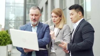 Team of business people employees in formal suits discussing a project together using a laptop and tablet standing near office building. Group partners mature person looking at computer screen outside