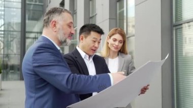 Diverse group business people outside office building, talking discussing new construction plan, team engineers and designers discuss holding and looking Technical drawing, drafting project
