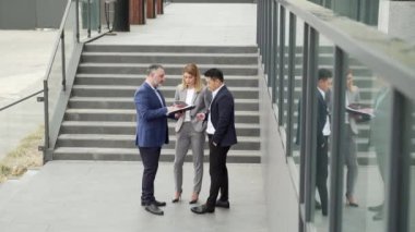 International business team meeting. mature man, Caucasian woman, Asian employee communicate . group of entrepreneurs in formal suits discuss a project document near office center on street. Outside