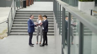 International business team meeting. mature man, Caucasian woman, Asian employee communicate . group of entrepreneurs in formal suits discuss a project document near office center on street. Outside