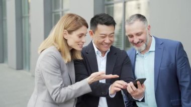 colleagues employees watch funny videos or look at photos, pictures memories looking at smartphone screen using mobile phone. group of friendly business people office workers having fun on breaks