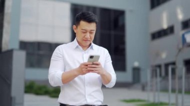 Asian office worker uses smartphone while standing outside. Handsome young business man employee chatting and browsing web using phone outdoors on city street Happy male texting smiling. downtown asia