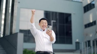 Asian business man employee entrepreneur looking phone smartphone with shouts win gesture, read good news, investment, profit growth. Happy male investor rejoices celebrate victory won outside office