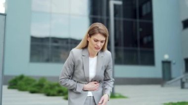 Business woman employee standing on city street with severe abdominal pain, upset stomach, diarrhea, premenstrual syndrome or poisoning. Female suffering holding stomach outside, near office building
