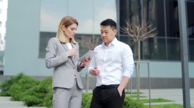 two employees partners stand in the street near the office business center, talking. team colleagues communicate discuss working project using tablet during break from outside. outdoors Downtown