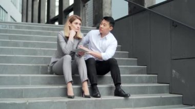 Asian male employee supports upset depressed female co-worker colleague. Sitting on the steps stairs outside an office building. young man comforting sad woman at work outdoors 
