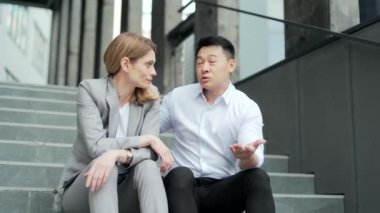 Asian male employee supports upset depressed female co-worker colleague. Sitting on the steps stairs outside an office building. young man comforting sad woman at work outdoors 