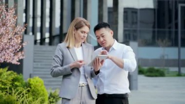 Two employees talking standing on a city street near office building. Using viewing Browsing a digital tablet. Business people Asian man and Caucasian woman discuss a project startup during a break